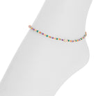 A delicate beaded anklet with small multicolored cylindrical beads and a central gold-toned oval charm, displayed on a white mannequin foot against a white background.