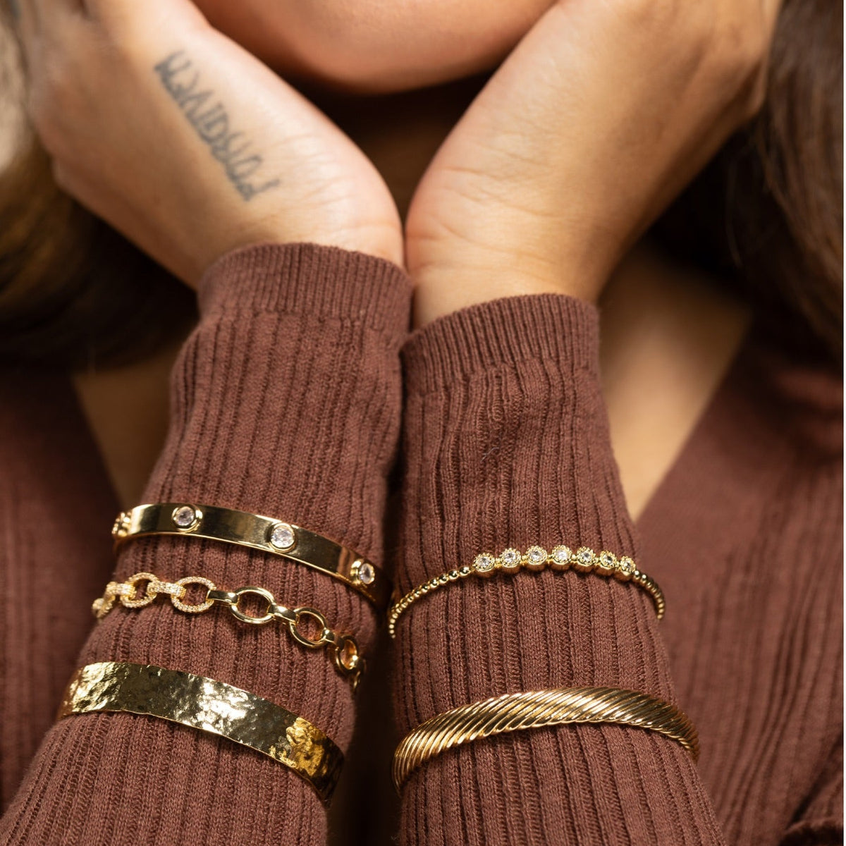 A close-up lifestyle shot of a model wearing multiple gold bracelets on both wrists, including a textured bangle, a chain bracelet, a diamond-studded bracelet, and a rope-style bangle, paired with a brown ribbed sweater.